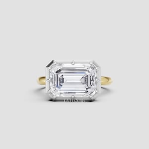 May include: An emerald-cut diamond ring with a white gold setting and a yellow gold band. The ring features a large, rectangular diamond with a clear, sparkling appearance. The ring is set against a neutral background.