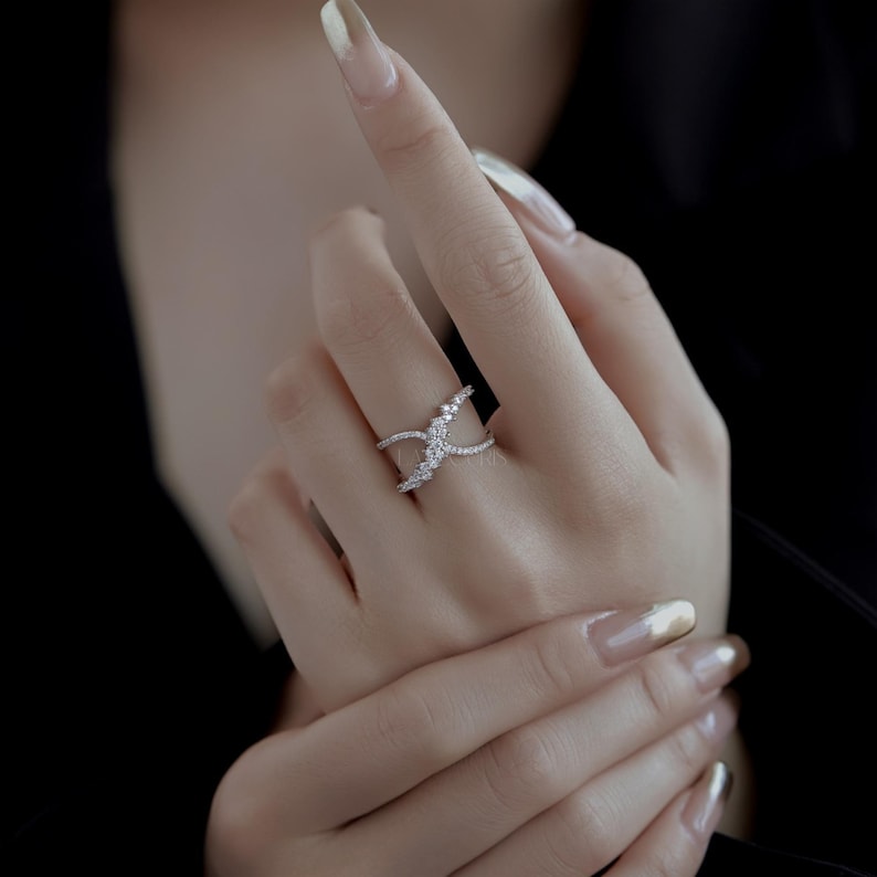 May include: A silver-toned ring with a unique, open design. The ring features a crisscross pattern with small, sparkling stones. The stones are clustered in the center and along the ring's bands, creating a dazzling effect. The ring is worn on a finger.