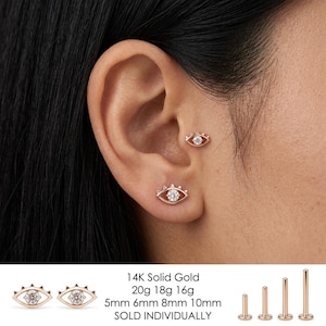 May include: A 14K solid gold eye-shaped stud earring featuring a central clear gemstone. The jewellery is available in 20g, 18g, and 16g gauges with post lengths of 5mm, 6mm, 8mm, and 10mm. The design includes a flat back post for secure placement.