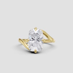 Puede incluir: Un anillo de compromiso dorado con una gema clara, grande y rectangular. La banda tiene un diseño único y ondulado. El anillo se encuentra sobre un fondo gris claro.
