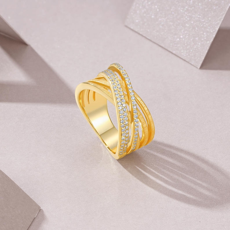 May include: A gold-colored ring with an interwoven design. The ring features multiple bands, some adorned with small, sparkling stones. The ring is set against a soft, neutral background, highlighting its intricate details and elegant form.
