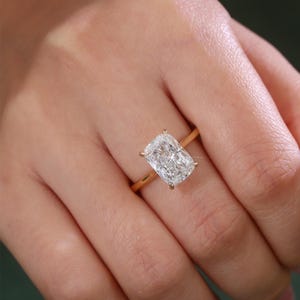 May include: A gold ring featuring a large, rectangular cushion-cut clear gemstone set in a four-prong setting. The band is a simple, polished gold metal, providing a clean and classic look that highlights the brilliance of the central stone.