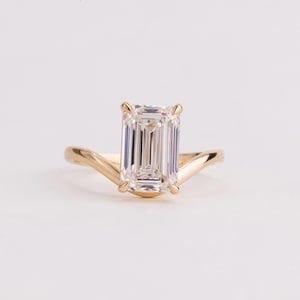 May include: A gold ring featuring a large, clear, emerald-cut gemstone set in a four-prong setting. The band has a distinct V-shaped curve that leads up to the central stone, creating a sleek and polished appearance with a warm metallic finish.