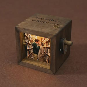 Può includere: Un cubo fotografico in legno con le parole "together, wherever" incise sopra. Il cubo presenta una foto di una coppia che si bacia all'interno di una biblioteca. Un piccolo anello d'oro è in cima al cubo.