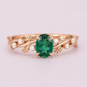 May include: Rose gold ring featuring an oval-cut emerald gemstone. The ring is adorned with leaf-like details and small, round, clear gemstones. The ring has the text "LA FLEURIS" engraved on the band.
