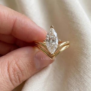 Könnte beinhalten: Ein Goldring mit einem transparenten Edelstein im Marquise-Schliff als Mittelstein, der von einer Vier-Krappen-Fassung gehalten wird. Die Ringschiene hat ein V-förmiges Chevron-Design, das sich um die Basis des Steins wölbt. LA ELOURIS steht auf dem Ring.
