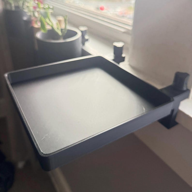 May include: A black plastic tray with a clip that attaches to a window sill. The tray is empty and appears to be designed for holding small items.