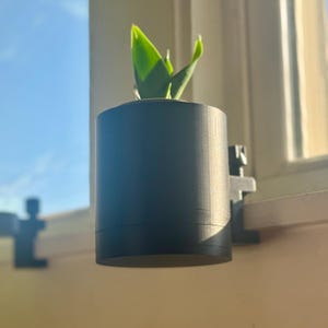 May include: A black plant pot with a small green plant inside is attached to a windowsill with a black clamp.