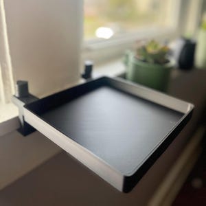 May include: A black tray with a white border is mounted on a window with a black clip. The tray is empty and ready to hold items.