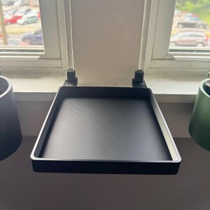 May include: Black metal windowsill plant shelf with two brackets. The shelf is mounted to the window frame and has a square shape.