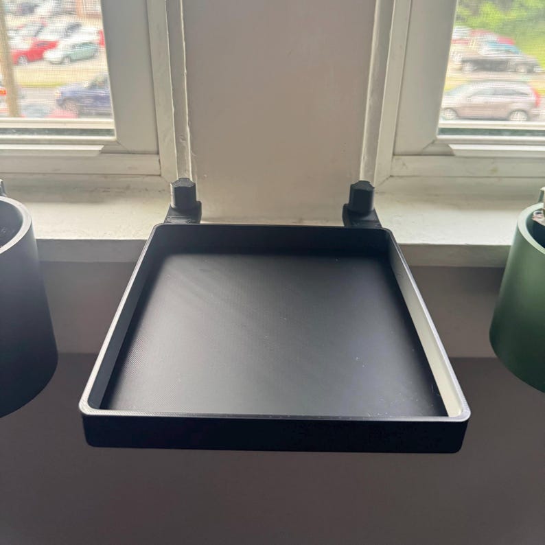 May include: Black metal windowsill plant shelf with two brackets. The shelf is mounted to the window frame and has a square shape.