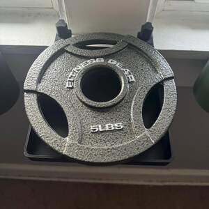 May include: A gray 5-pound weight plate with the text "Fitness Gear" printed on it. The plate is hanging on a black metal rack.