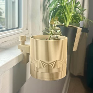 May include: A beige planter with a geometric pattern is attached to a windowsill with a clamp. The planter is holding a small succulent plant.