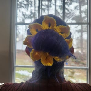 Può includere: Capelli di una donna acconciati con un girasole all'uncinetto giallo e capelli viola. Il girasole è posizionato sulla parte posteriore della testa, con i petali che incorniciano i capelli. I capelli sono acconciati in uno chignon.