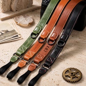 May include: Four camera straps in olive green, tan, light brown, and black leather. Each strap features metal D-rings, reinforced stitching, and black nylon loops at the ends. The straps are personalized with embossed text including Daniel, John Smith, LBA, and MLJ.