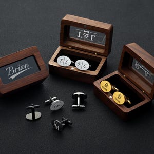 May include: Wooden boxes with personalized cufflinks. One box displays the name "Brian", while others contain silver and gold cufflinks with initials and names. The cufflinks are in various shapes and colors.