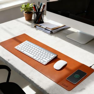 K&ouml;nnte beinhalten: Eine braune Leder-Schreibtischunterlage mit kabelloser Ladestation, silberner Tastatur und wei&szlig;er Maus. Auf der Unterlage sind die Initialen "C&D" eingepr&auml;gt. Ein Smartphone wird auf der Ladestation geladen. Ein Schreibtisch mit Pflanze, Stiften und Computermonitor im Hintergrund.