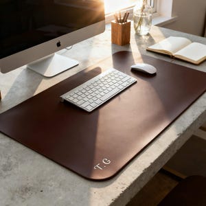 May include: A dark brown leather desk mat with rounded corners, a keyboard, and a computer mouse. The mat has the initials "T.G." in the corner. A computer monitor, a wooden pencil holder, and an open notebook are also visible.