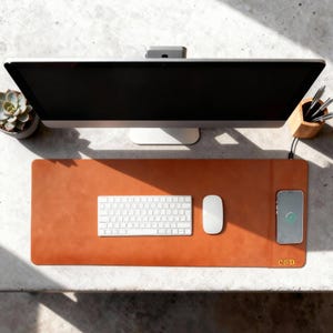 K&ouml;nnte beinhalten: Draufsicht auf einen Arbeitsplatz mit einer braunen Leder-Schreibtischunterlage. Dazu geh&ouml;ren eine wei&szlig;e Tastatur, eine Maus und ein Smartphone. Ein Computermonitor, eine Pflanze und ein Stifthalter sind ebenfalls zu sehen. Die Unterlage hat die Initialen "C&D".