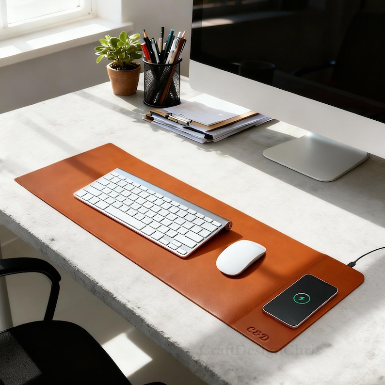 K&ouml;nnte beinhalten: Eine braune Leder-Schreibtischunterlage mit kabelloser Ladestation, silberner Tastatur und wei&szlig;er Maus. Auf der Unterlage sind die Initialen "C&D" eingepr&auml;gt. Ein Smartphone wird auf der Ladestation geladen. Ein Schreibtisch mit Pflanze, Stiften und Computermonitor im Hintergrund.