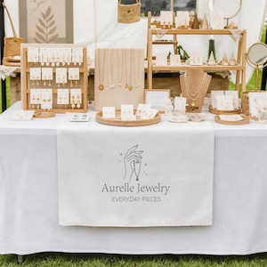 Op de afbeelding: Een witte tafeldisplay met diverse goudkleurige kettingen en oorbellen op standaards en kaarten. Een centrale witte tafelloper toont de tekst Aurelle Jewelry EVERYDAY PIECES met een lijntekening van een hand die een kettinghanger vasthoudt.