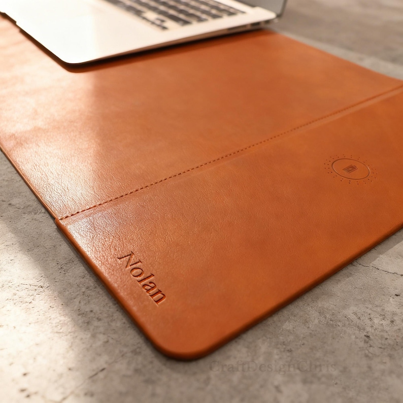 K&ouml;nnte beinhalten: Eine braune Leder-Schreibtischunterlage mit dem gepr&auml;gten Namen "Nolan" in der unteren linken Ecke. Die Unterlage hat eine Naht und ein kreisf&ouml;rmiges Design auf der rechten Seite. Ein Laptop liegt auf der Unterlage.