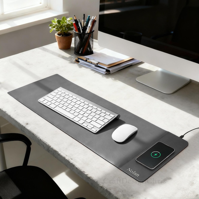 K&ouml;nnte beinhalten: Eine graue Schreibtischunterlage mit kabellosem Ladepad, Tastatur und Maus. Ein Smartphone wird auf dem Pad geladen. Auf der Matte steht der Name "Nolan". Ein Blumentopf, Bleistifte und ein Computer befinden sich ebenfalls auf dem Schreibtisch.