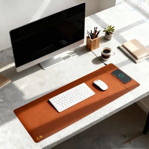 K&ouml;nnte beinhalten: Eine braune Leder-Schreibtischunterlage mit kabellosem Ladepad, Tastatur und Maus. Ein Computermonitor, eine Kaffeetasse und eine kleine Pflanze befinden sich ebenfalls auf dem Schreibtisch. Die Matte hat die Initialen "E.K.".