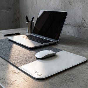 Op de afbeelding: Een moderne werkplek met een laptop, een witte muis en een gepersonaliseerde muismat. De muismat is gemaakt van grijs vilt met een wit leren gedeelte, met de initialen "T.G.". Een pen en een pennenhouder zijn ook zichtbaar.