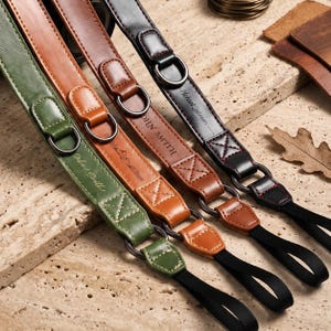 May include: Four leather camera straps in green, light brown, medium brown, and black. Each strap features metal D-rings, reinforced cross-stitching, and black nylon webbing loops at the ends. The straps are personalized with engraved names.