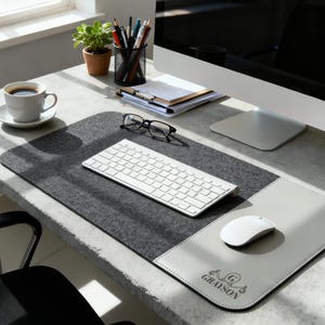 May include: A contemporary desk arrangement with a grey felt desk mat, a white keyboard, and a white mouse. A white coffee cup, a small potted plant, and stationery are also present. The desk mat has a light grey leather mouse pad with the name "GRAYSON".
