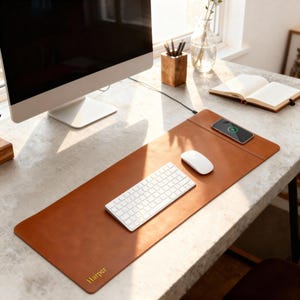 Pode incluir: Um tapete de mesa de couro castanho com um carregador sem fios, teclado e rato. O tapete tem o nome "Harper" impresso em dourado. Um monitor de computador, um porta-lápis de madeira e um caderno também são visíveis.