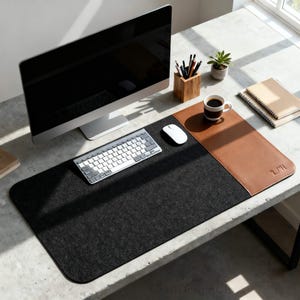 Op de afbeelding: Een moderne bureau-opstelling met een grote computermonitor, een draadloos toetsenbord en een muis. Een zwarte vilten bureaumat met een bruin lederen gedeelte bevat een kopje koffie, pennen en een kleine plant. Het bureau is van beton.