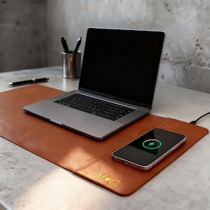 K&ouml;nnte beinhalten: Eine braune Leder-Schreibtischunterlage mit integrierter kabelloser Ladestation. Ein silberner Laptop und ein Smartphone liegen darauf. Das Smartphone wird geladen. Die Unterlage hat die Initialen "M C" und ein Herzsymbol.