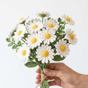 Könnte beinhalten: Ein Strauß aus gehäkelten Gänseblümchen mit weißen Blütenblättern und leuchtend gelben Mitten. Die Blumen sind an grünen Stielen mit kleinen grünen Blättern befestigt, die an der Basis mit einer einfachen Schnur zusammengebunden sind.