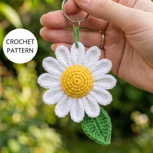 Può includere: Un portachiavi a forma di margherita all'uncinetto con un centro giallo brillante, petali bianchi e una singola foglia verde attaccata a un anello in metallo argentato. Il testo CROCHET PATTERN è stampato in nero su un cerchio bianco in alto a sinistra.