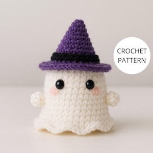 May include: A white crocheted ghost figurine wearing a purple witch hat with a black band. The ghost has two black bead eyes and small pink blush marks on its cheeks. A white circular graphic on the right side contains the text CROCHET PATTERN.