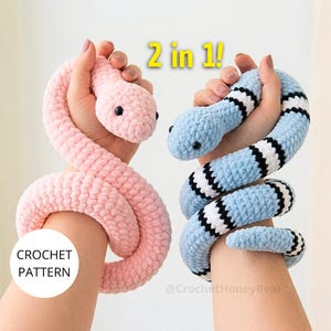 Puede incluir: Dos serpientes de ganchillo de felpa, una de color rosa claro sólido y otra a rayas de color azul claro, blanco y negro. El texto 2 in 1! aparece en la parte superior y el texto CROCHET PATTERN es visible en un círculo blanco a la izquierda.