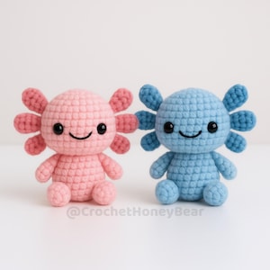 May include: Two handmade crochet axolotl plushies. One is pink with darker pink gills, the other is blue. Both have black eyes and smiling faces. The axolotls are sitting on a white surface.
