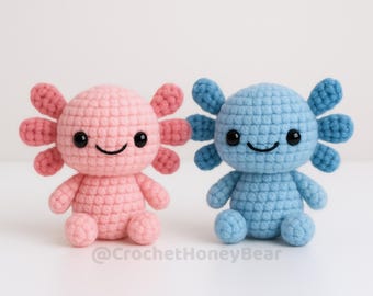 Op de afbeelding: Twee handgemaakte gehaakte axolotl-knuffels. Eén is roze met donkerroze kieuwen, de andere is blauw. Beide hebben zwarte ogen en lachende gezichten. De axolotls zitten op een wit oppervlak.