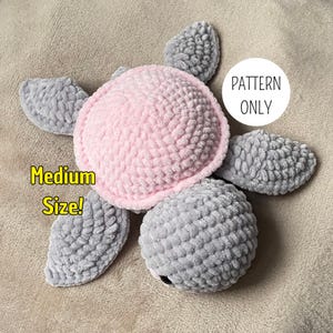 Peut inclure: Une peluche tortue au crochet avec une carapace rose tendre et une tête ainsi que des nageoires grises. Le texte sur l'image indique "Medium Size!" et "PATTERN ONLY". La peluche est faite d'un fil texturé et épais au corps arrondi.