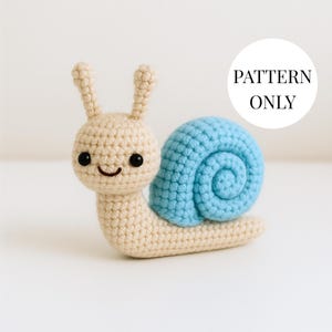 May include: A crocheted snail toy with a light beige body and a light blue shell. The snail has black eyes and a smiling mouth. The text "PATTERN ONLY" is in a white circle in the background.