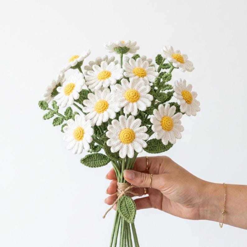 May include: A bouquet of crocheted daisy flowers featuring white petals and bright yellow centers. The flowers are attached to green stems with small green leaves, all tied together with a simple piece of twine at the base of the stems.