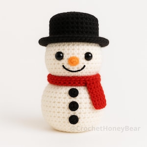 Puede incluir: Muñeco de nieve tejido a ganchillo con sombrero de copa negro y bufanda roja. Tiene ojos negros, nariz naranja y boca sonriente. El muñeco de nieve tiene tres botones negros. El texto "CrochetHoneyBear" está en la parte inferior.