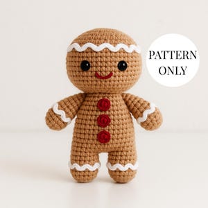 May include: A crocheted gingerbread man doll with a smiling face, black eyes, and a red mouth. The doll has white icing details and red button accents. The text "PATTERN ONLY" is in a white circle.