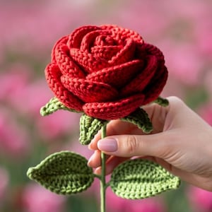 May include: A handcrafted crochet rose featuring deep red yarn petals arranged in a layered, blooming pattern. The flower is attached to a thin green stem with two textured green leaves extending from the sides, all created with detailed yarn stitching.