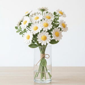 May include: A bouquet of crocheted daisy flowers with white petals and yellow centers, featuring green crocheted leaves and stems. The flowers are arranged in a clear glass cylindrical vase, tied together with a thin piece of brown twine.