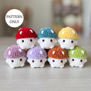 May include: A collection of eight small crocheted mushroom plushies with white stems and caps in red, blue, yellow, purple, lavender, orange, and green, each featuring white spots and black eyes. A white circular label reads PATTERN ONLY.
