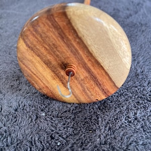 May include: A drop spindle featuring a circular wooden whorl with a polished finish showing natural light and dark brown wood grain patterns. A thin metal hook is centered on the top surface, extending from a small, cylindrical wooden bead.