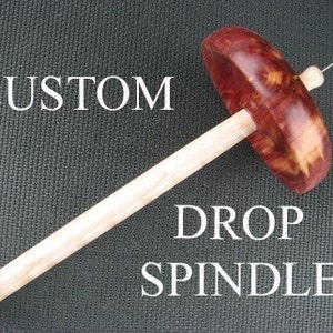 May include: A custom drop spindle with a wooden shaft and a red and brown wood whorl. The whorl has a unique grain pattern. The spindle is labeled "CUSTOM DROP SPINDLE".